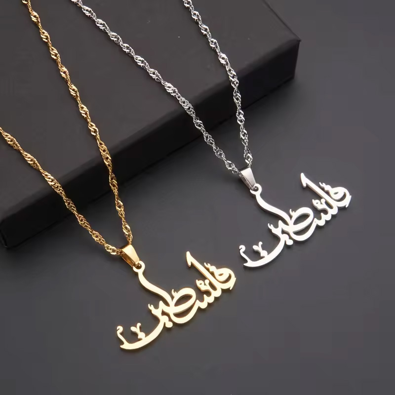 Two necklaces with Arabic script pendants, one in gold and one in silver, displayed on a dark background. Keywords: necklace.