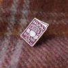 Burgundy enamel pin with "Passport Scotland Alba" text and a thistle design, placed on a plaid fabric.