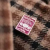 Burgundy enamel pin with "Passport Scotland Alba" text and a thistle design, placed on a plaid fabric close-up.