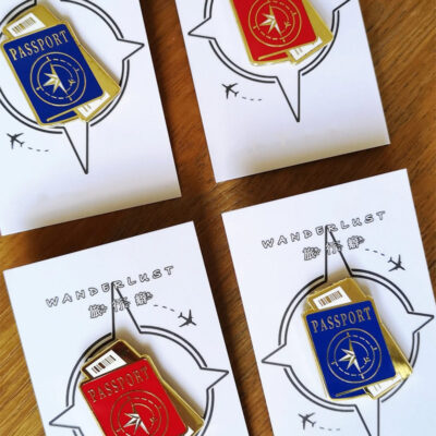 Four enamel pins in the shape of passports, two red and two blue, with gold details, displayed on branded cards.