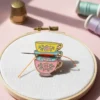 A needle and thread on a table beside a tea cup and an enamel needle minder, creating a cozy crafting scene.