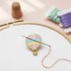Hot air balloon enamel pin on a cross stitch fabric, serving as a needle minder for sewing enthusiasts.