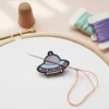 Cross stitch featuring a purple and white alien, designed as an enamel needle minder for crafting enthusiasts.