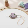 A purple alien enamel pin attached to a needle and thread, designed as a needle minder for sewing enthusiasts.