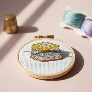 Small embroidered enamel pin featuring a stack of colorful books, designed as a needle minder for sewing enthusiasts.