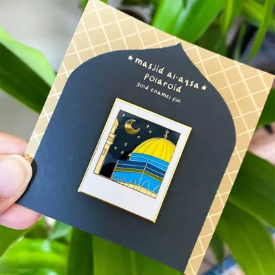 Custom enamel pin featuring the Al-Aqsa Mosque with a night sky background, held by a person against a green leaf backdrop.