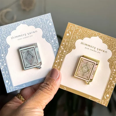 A pair of enamel pins depicting the Quran, one in silver and one in gold, with intricate designs, held against ornate backing cards.