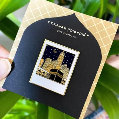 A gold enamel pin featuring an illustration of the Kaabah and surrounding buildings under a night sky, presented on a black card with the text "Kaabah Polaroid."