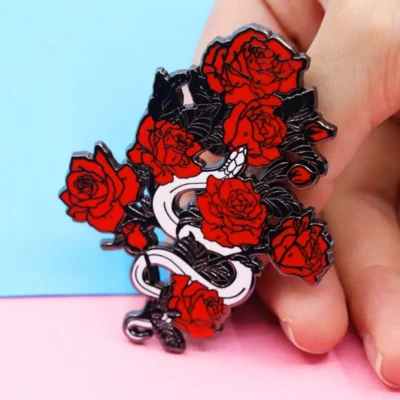 A hand delicately holds a red rose enamel pin, showcasing its vibrant color and intricate design against a neutral background.