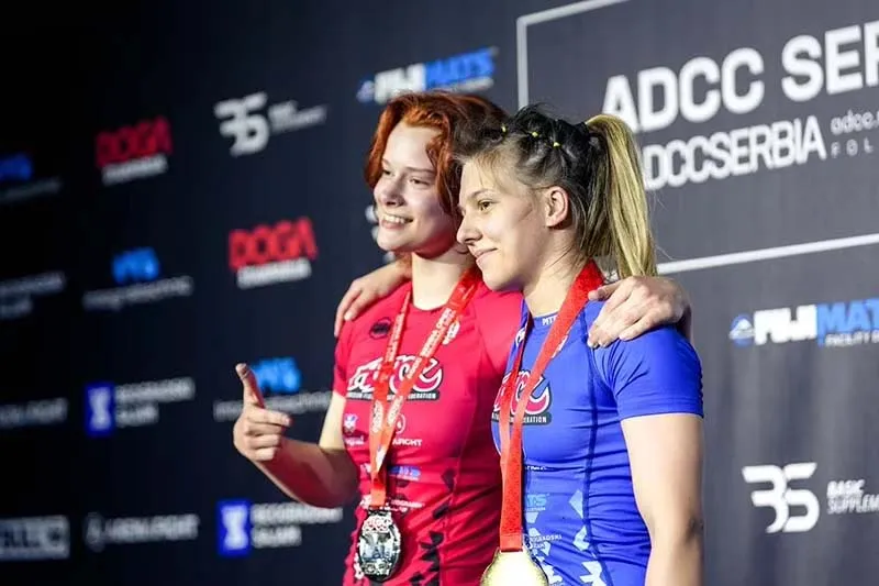 Two women proudly stand on a podium, displaying their ADCC medals, celebrating their achievements in a competitive event.