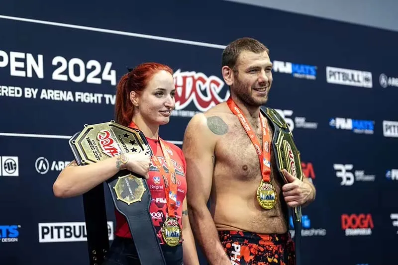 A man and woman proudly display their ADCC medals and championship belt while standing on stage during the award ceremony.