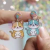 Une personne présentant des boucles d'oreilles en émail souple personnalisées, ornées d'un chat enjoué.