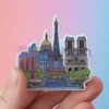 Una mano sujeta una pegatina con la Torre Eiffel y varios edificios, para mostrar imanes de nevera personalizados para viajes.
