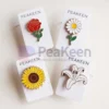 Une image présentant quatre magnifiques épingles à fleurs en émail mou : une rose rouge vibrante, une marguerite blanche délicate avec un centre jaune, un tournesol jaune vif avec un centre marron riche, et un élégant lys blanc. Chaque épingle est montée sur sa propre carte en papier lisse, avec le nom de la marque "PeaKeen" imprimé en évidence dans la partie supérieure. La combinaison de couleurs vives et la finition émaillée brillante mettent en valeur les détails complexes de chaque fleur, tandis que la présentation uniforme sur des cartes de marque ajoute une touche professionnelle et cohérente. Parfait pour les cadeaux ou la vente au détail.