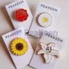 Une image présentant quatre magnifiques épingles à fleurs en émail mou : une rose rouge vibrante, une marguerite blanche délicate avec un centre jaune, un tournesol jaune vif avec un centre marron riche, et un élégant lys blanc. Chaque épingle est montée sur sa propre carte en papier lisse, avec le nom de la marque "PeaKeen" imprimé en évidence dans la partie supérieure. La combinaison de couleurs vives et la finition émaillée brillante mettent en valeur les détails complexes de chaque fleur, tandis que la présentation uniforme sur des cartes de marque ajoute une touche professionnelle et cohérente. Parfait pour les cadeaux ou la vente au détail.