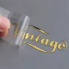 Primer plano de una persona agarrando una lámina dorada con la palabra "vintage" impresa en una pegatina metálica, que resalta una estética clásica.