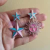 Pendientes de esmalte personalizados con un diseño de dos estrellas y un sol, adornados con vibrantes detalles de esmalte azul.