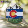Pin de la bandera de Colorado con montañas y árboles, símbolo de la belleza natural y el orgullo del estado.