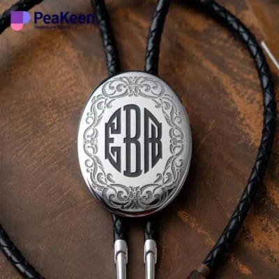 A black leather lanyard featuring a silver oval with a monogram, designed for a stylish bolo tie.