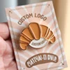 A person holding a croissant-shaped pin featuring the words "custom logo" prominently displayed.