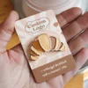 Close-up of a hand holding a brown and white enamel pin in the shape of a croissant.