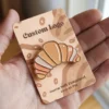 A hand presents a small enamel pin designed as a croissant, emphasizing its colorful and artistic features.