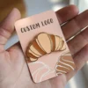 A hand holding a small enamel pin shaped like a croissant, featuring the words "custom logo" prominently displayed.