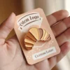 A person holds a small enamel pin featuring a croissant design with the word "custom logo" in stylish lettering.