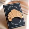 A pin featuring a croissant design with the words "Custom Logo" prominently displayed.