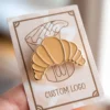 A card showcasing a gold enamel pin in the shape of a croissant.