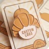 Enamel pin designed as a croissant, highlighting a unique and creative logo for branding or personal use.