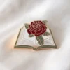 Роза лежит на раскрытой книге, рядом с золотой эмалевой булавкой, которая символизирует любовь к чтению.