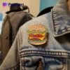 Einzelperson in Jeansjacke mit verspielter Hamburger-Anstecknadel, die ein einzigartiges Modestatement setzt.