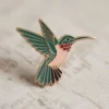Un vibrante alfiler de esmalte de colibrí sobre fondo blanco, que resalta su delicado diseño y sus colores.