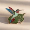 Un vibrante alfiler de esmalte de colibrí que descansa sobre una hoja verde, mostrando intrincados detalles y colores.