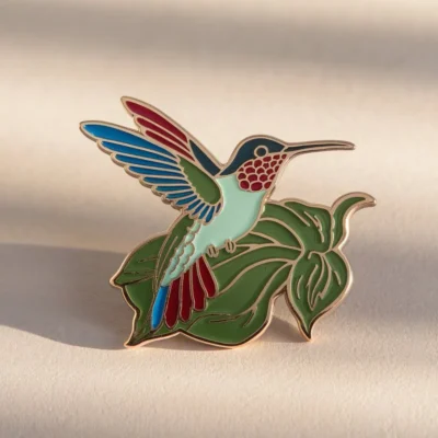 A vibrant hummingbird enamel pin resting on a green leaf, showcasing intricate details and colors.