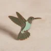 Alfiler de esmalte con forma de colibrí sobre un fondo blanco limpio, que muestra detalles intrincados y colores vibrantes.