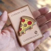Una mano sujeta un pin con temática de pizza pegado a una tarjeta de felicitación, mostrando un diseño divertido y juguetón.