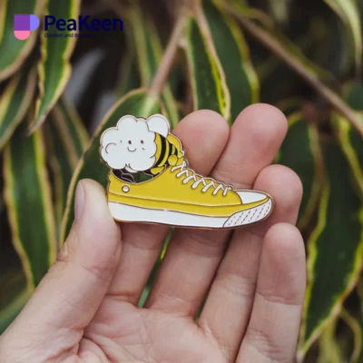 Una mano presenta un zapato amarillo con un alfiler de esmalte de abeja, destacando una creativa mezcla de moda y capricho.