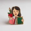 A pin featuring a woman holding an apple in one hand and a book in the other, symbolizing a teacher's role.