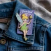 A Tinkerbell enamel pin rests on a blue jean jacket, showcasing a whimsical fairy design.