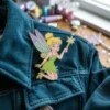 A Tinkerbell enamel pin featuring a fairy, resting on a blue jacket.