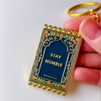 Close-up of a hand gripping a prayer mat keychain inscribed with "stay humble," symbolizing a reminder for humility.
