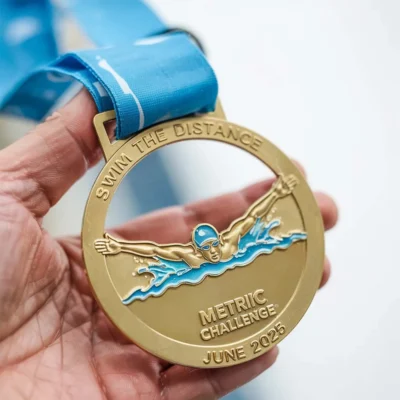 A person proudly holds a gold enamel swimming medal inscribed with "metric challenge."