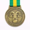 A round gold Premier Rugby League Grand Final 2026 medal featuring a ram’s head above a rugby ball, with a textured background and a green and yellow ribbon.