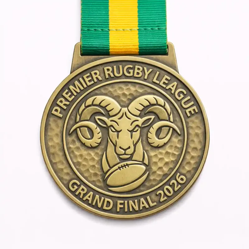 A round gold Premier Rugby League Grand Final 2026 medal featuring a ram’s head above a rugby ball, with a textured background and a green and yellow ribbon.