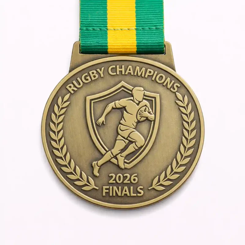 A gold Rugby Champions 2026 Finals medal featuring a running rugby player inside a shield, surrounded by laurel wreaths, with a green and yellow ribbon.