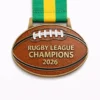 An oval-shaped Rugby League Champions 2026 medal designed like a rugby ball, featuring textured brown enamel, gold detailing, white laces at the top, and a green and yellow ribbon.