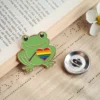 Enamel frog pin with a rainbow heart beside an open book on a wooden surface.