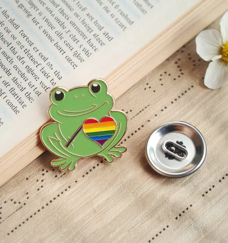 Enamel frog pin with a rainbow heart beside an open book on a wooden surface.